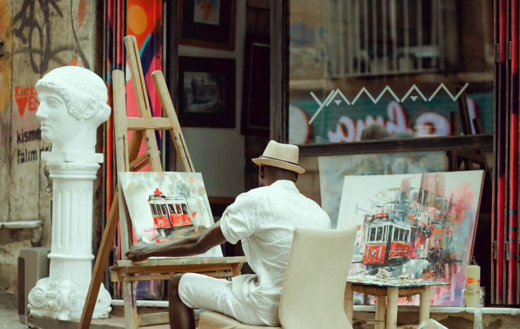 Pintor sentado numa calçada pintando uma tela em frente uma loja