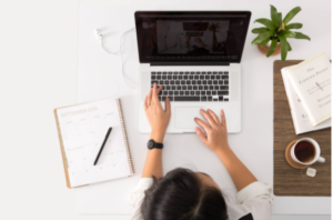 Foto vista de cima de uma mulher com notebook aberto e cadernos sobre a mesa